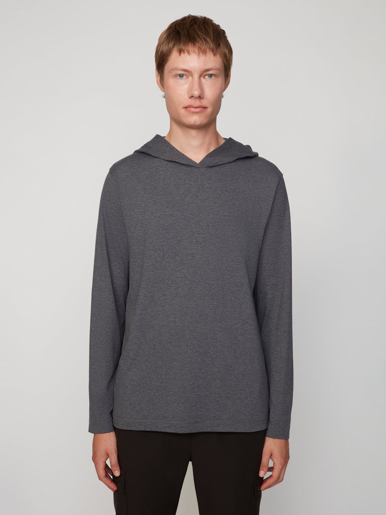 A person with short light brown hair stands against a plain background, wearing TECH APPAREL’s MEN’S LONG SLEEVE HOODIE IN BRUSHED SPACE-DYED JERSEY and black pants—a versatile, stylish layer for any occasion.