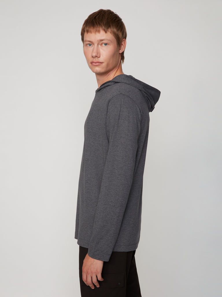 A young man with short light brown hair stands sideways, looking at the camera. He wears the TECH APPAREL MEN’S LONG SLEEVE HOODIE IN BRUSHED SPACE-DYED JERSEY and black pants against a plain light background.