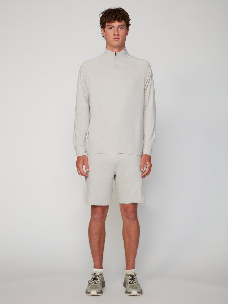 A young man faces forward against a plain light background, wearing TECH APPAREL’s MEN’S 1/4 ZIP MOCK NECK LONG SLEEVE IN BRUSHED SPACE-DYED JERSEY with matching shorts and gray sneakers.