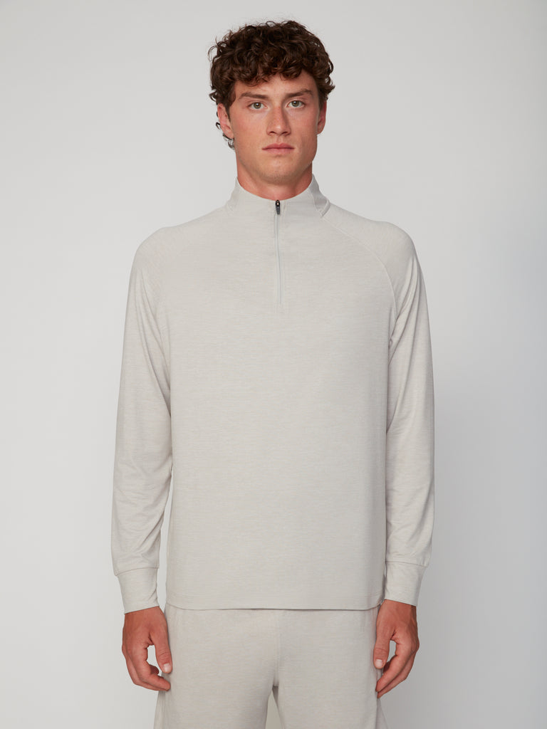 A young man with curly brown hair wears the TECH APPAREL MEN’S 1/4 ZIP MOCK NECK LONG SLEEVE IN BRUSHED SPACE-DYED JERSEY in light beige with matching pants, standing against a plain light gray background, looking at the camera.