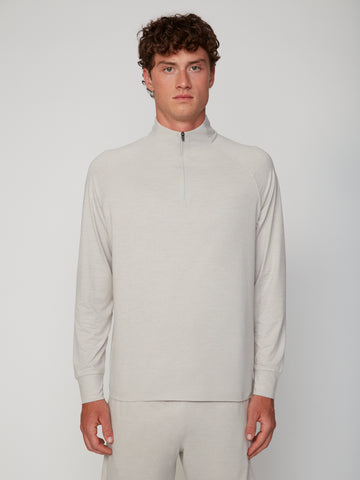 A young man with curly brown hair wears the TECH APPAREL MEN’S 1/4 ZIP MOCK NECK LONG SLEEVE IN BRUSHED SPACE-DYED JERSEY in light beige with matching pants, standing against a plain light gray background, looking at the camera.
