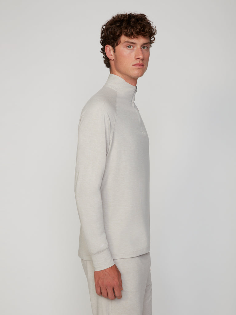 A young man with curly brown hair stands in profile, wearing the TECH APPAREL MEN’S 1/4 ZIP MOCK NECK LONG SLEEVE in brushed space-dyed jersey, paired with matching pants, set against a plain light background.