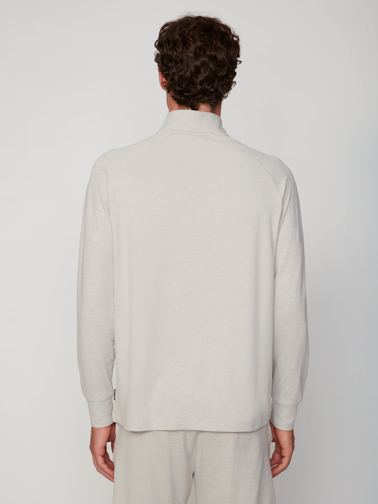 A person with short curly brown hair stands with their back to the camera, wearing the TECH APPAREL MEN’S 1/4 ZIP MOCK NECK LONG SLEEVE in brushed space-dyed jersey against a plain light background.