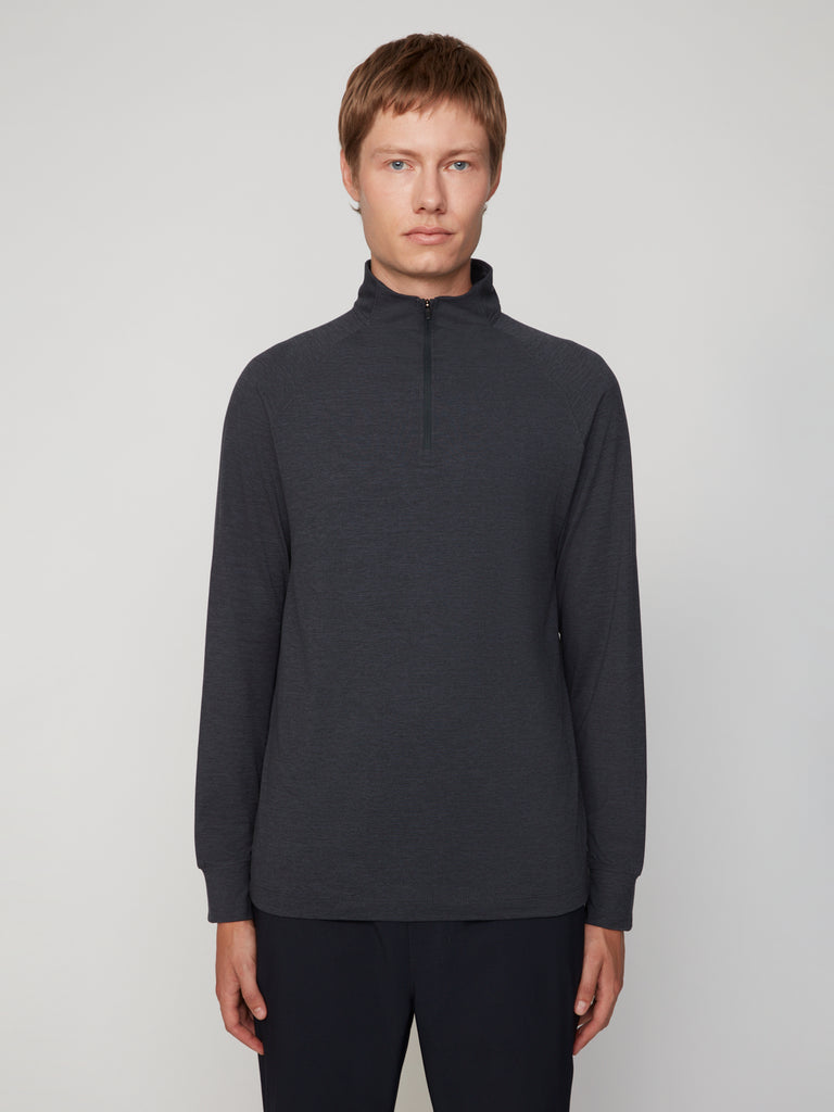 A man with short light brown hair wears the TECH APPAREL MEN’S 1/4 ZIP MOCK NECK LONG SLEEVE IN BRUSHED SPACE-DYED JERSEY in dark gray and dark pants, standing against a plain light gray background, facing forward neutrally.