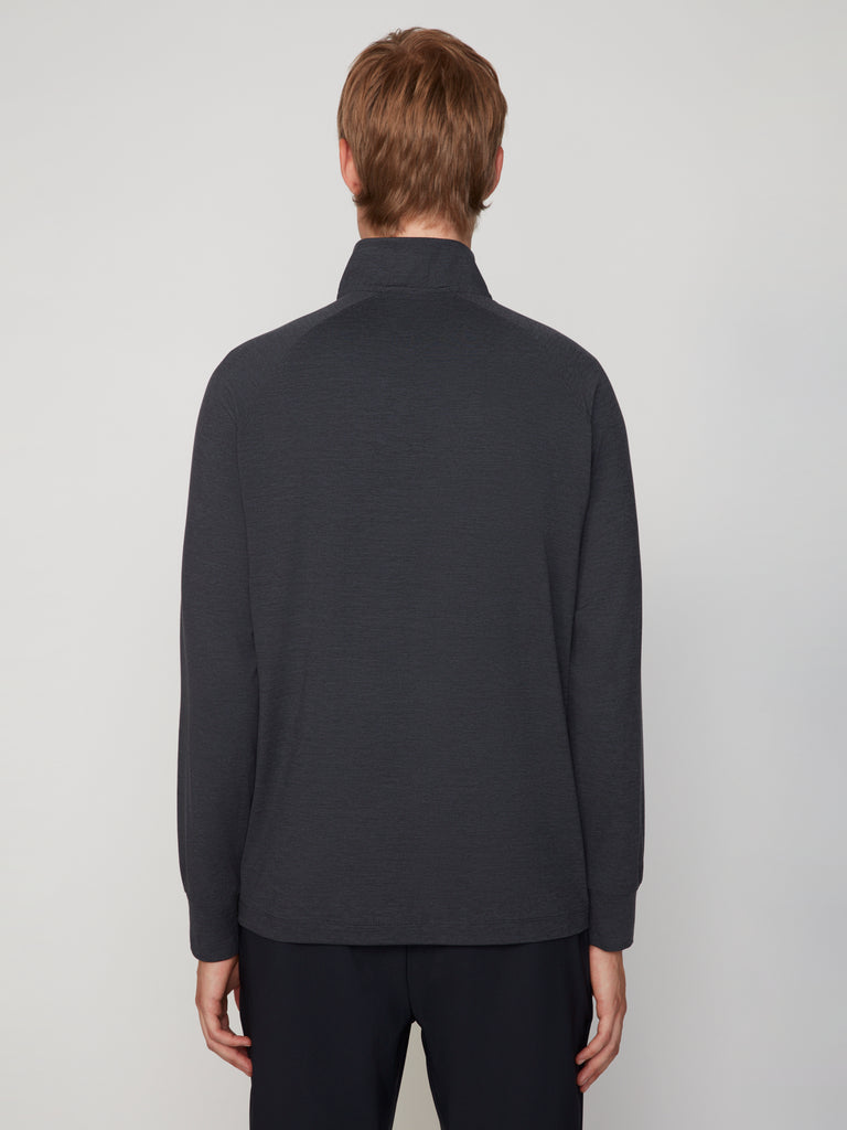 A person with short light brown hair faces away from the camera, wearing TECH APPARELs MEN’S 1/4 ZIP MOCK NECK LONG SLEEVE IN BRUSHED SPACE-DYED JERSEY and dark pants against a plain light background, highlighting performance-driven comfort.