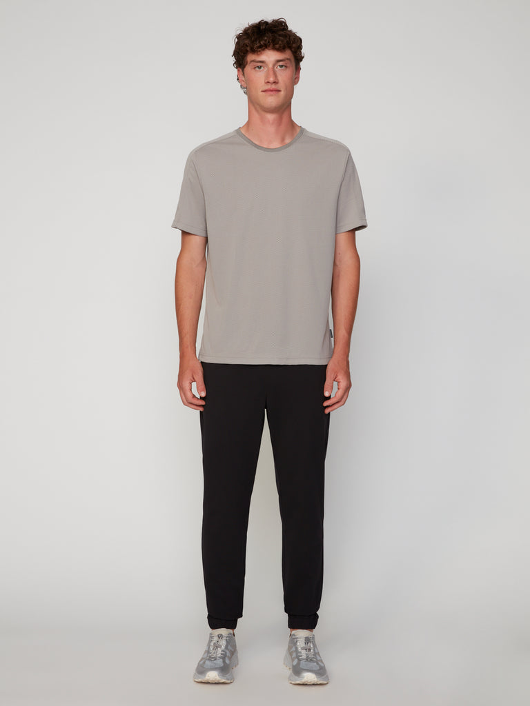 A young man with short, curly hair stands facing forward against a light background, wearing a TECH APPAREL MEN’S EYELET JACQUARD CREW NECK T-SHIRT in light gray, black jogger pants, and gray sneakers with his arms at his sides.