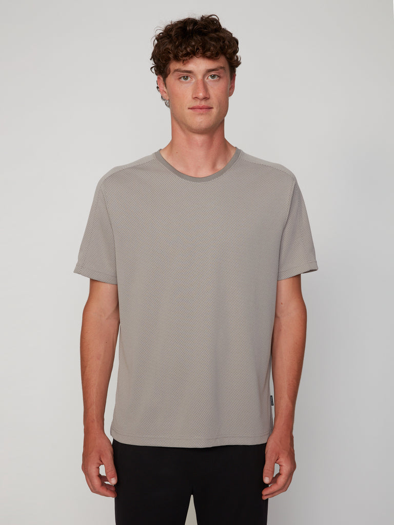 A young man with curly brown hair stands facing forward against a plain white background, wearing a light gray TECH APPAREL MEN’S EYELET JACQUARD CREW NECK T-SHIRT and black pants.