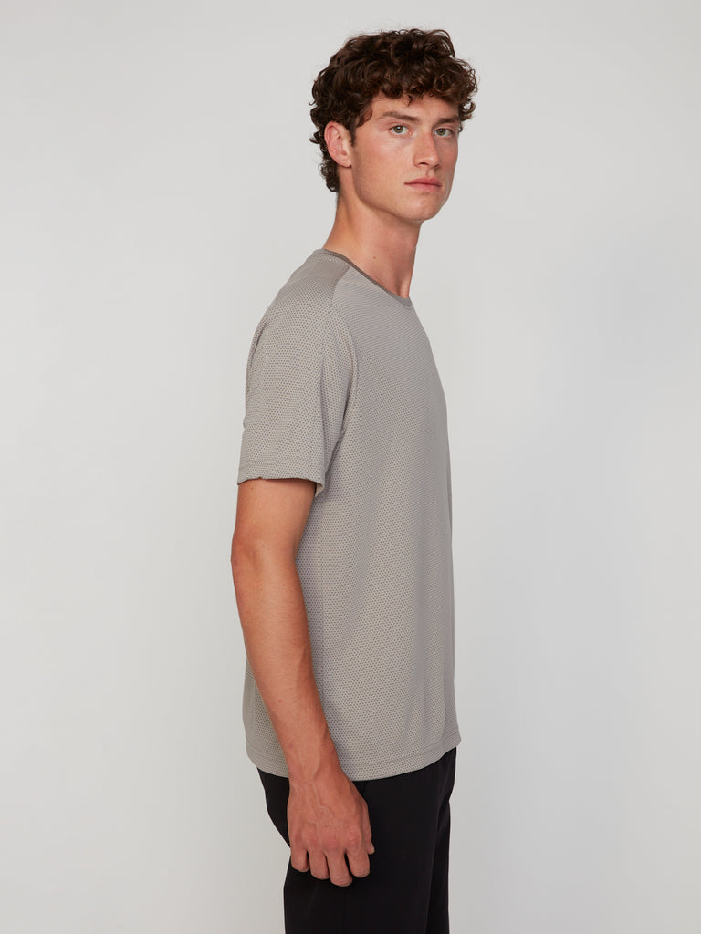 A young man with curly hair stands sideways, looking at the camera. He wears a light gray TECH APPAREL MEN’S EYELET JACQUARD CREW NECK T-SHIRT and black pants against a plain white background.