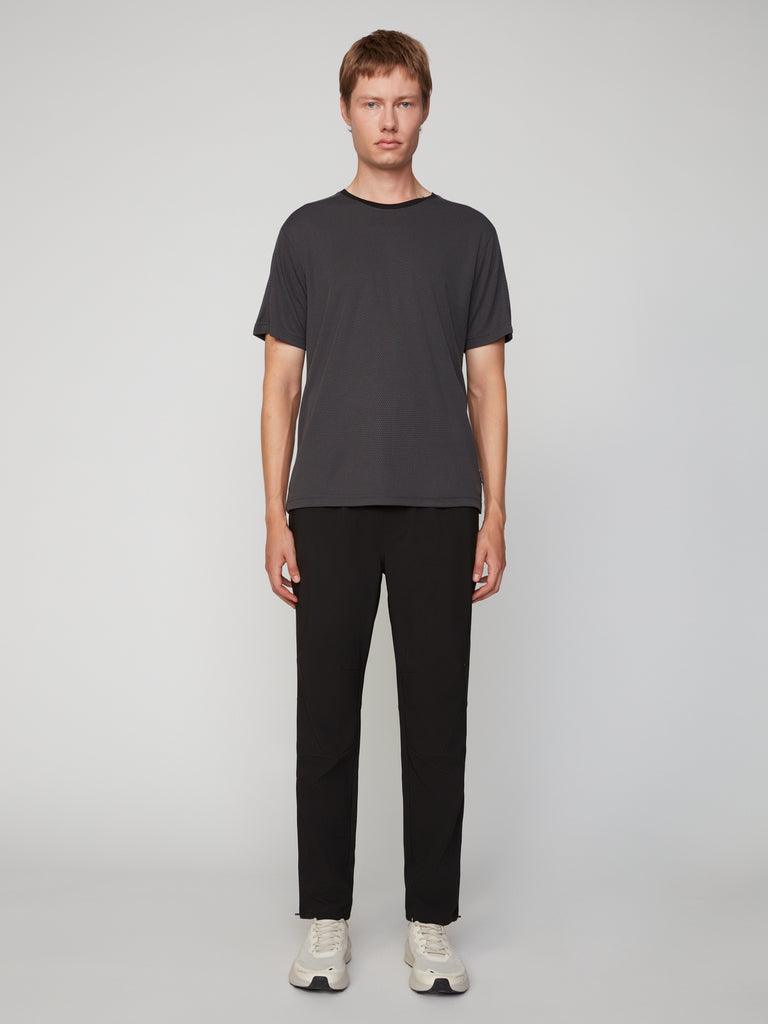 A person with short hair faces forward against a plain light background, wearing TECH APPAREL’s MEN’S EYELET JACQUARD CREW NECK T-SHIRT in dark gray, black pants, and white sneakers.