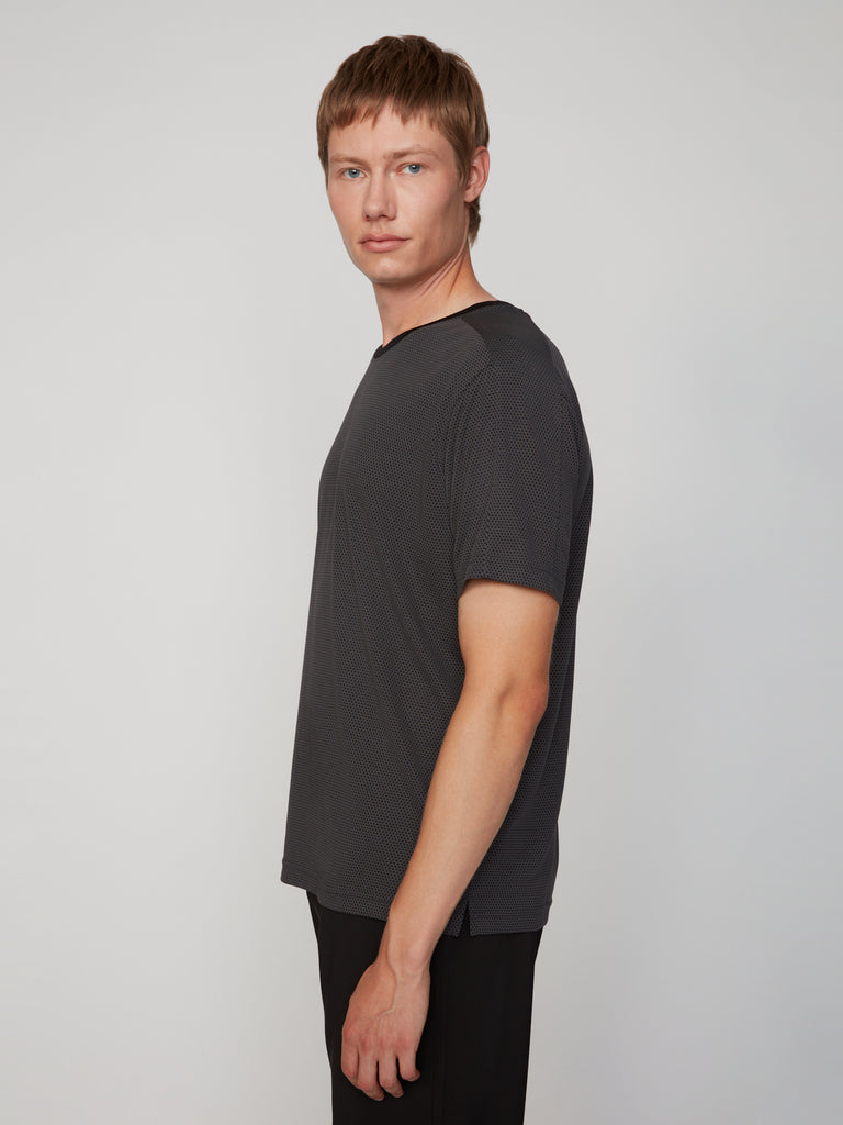 A person with short light brown hair wears the TECH APPAREL MEN’S EYELET JACQUARD CREW NECK T-SHIRT and dark pants, standing sideways and looking at the camera against a plain light background.