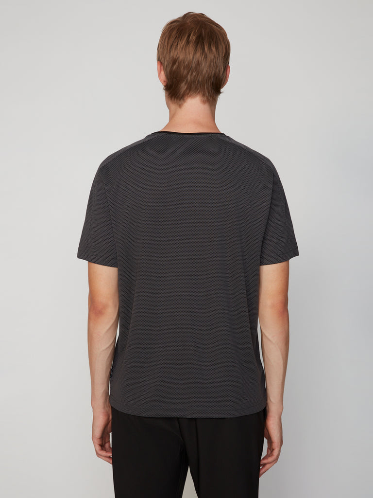 A person with light brown hair stands with their back to the camera, wearing TECH APPAREL’s MEN’S EYELET JACQUARD CREW NECK T-SHIRT in dark gray and black pants against a plain light gray background.