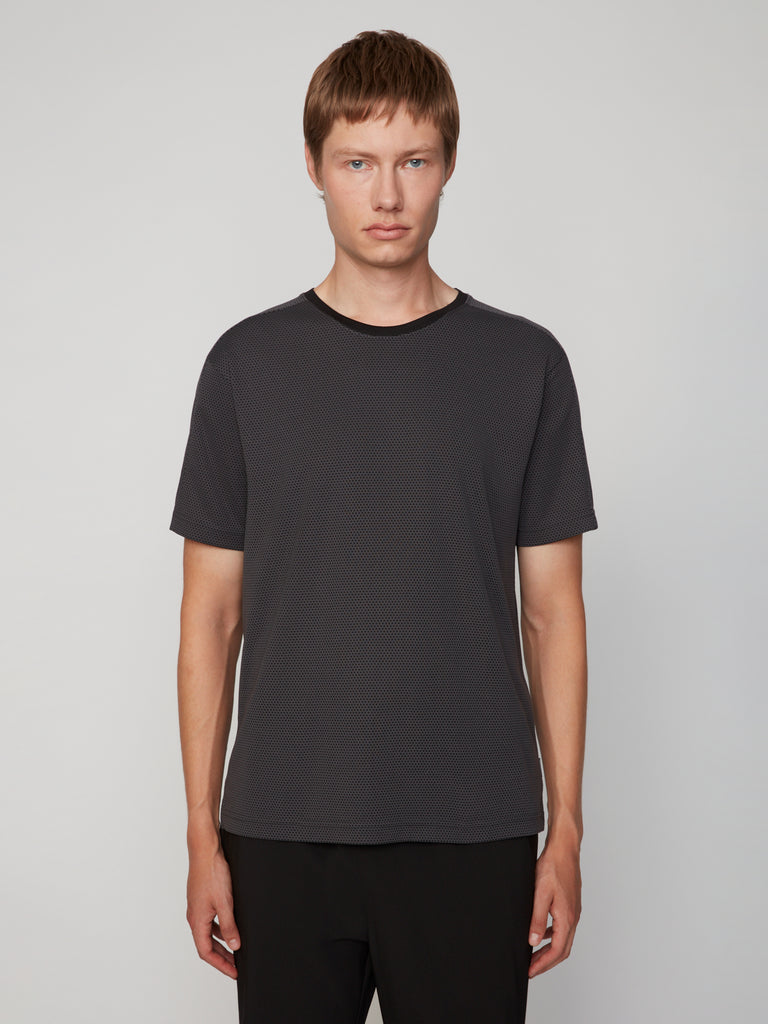 A young person with short light brown hair wears the TECH APPAREL MEN’S EYELET JACQUARD CREW NECK T-SHIRT in dark gray and black pants, standing against a plain light gray background and looking at the camera with a neutral expression.