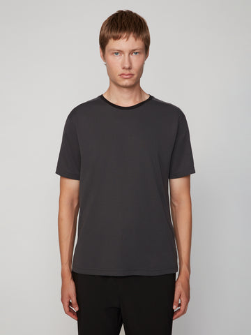 A young person with short light brown hair wears the TECH APPAREL MEN’S EYELET JACQUARD CREW NECK T-SHIRT in dark gray and black pants, standing against a plain light gray background and looking at the camera with a neutral expression.