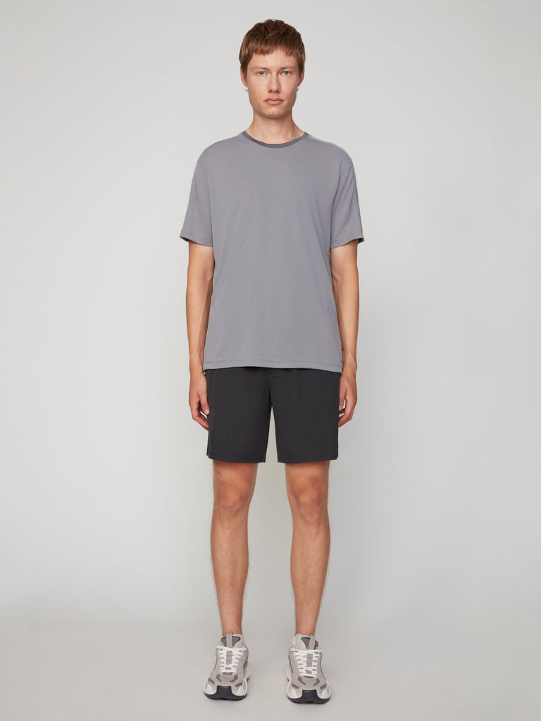A person stands facing forward against a plain light background, wearing the TECH APPAREL MEN’S EYELET JACQUARD CREW NECK T-SHIRT in gray, black shorts, and gray athletic shoes. Their arms are relaxed at their sides and they have short brown hair.