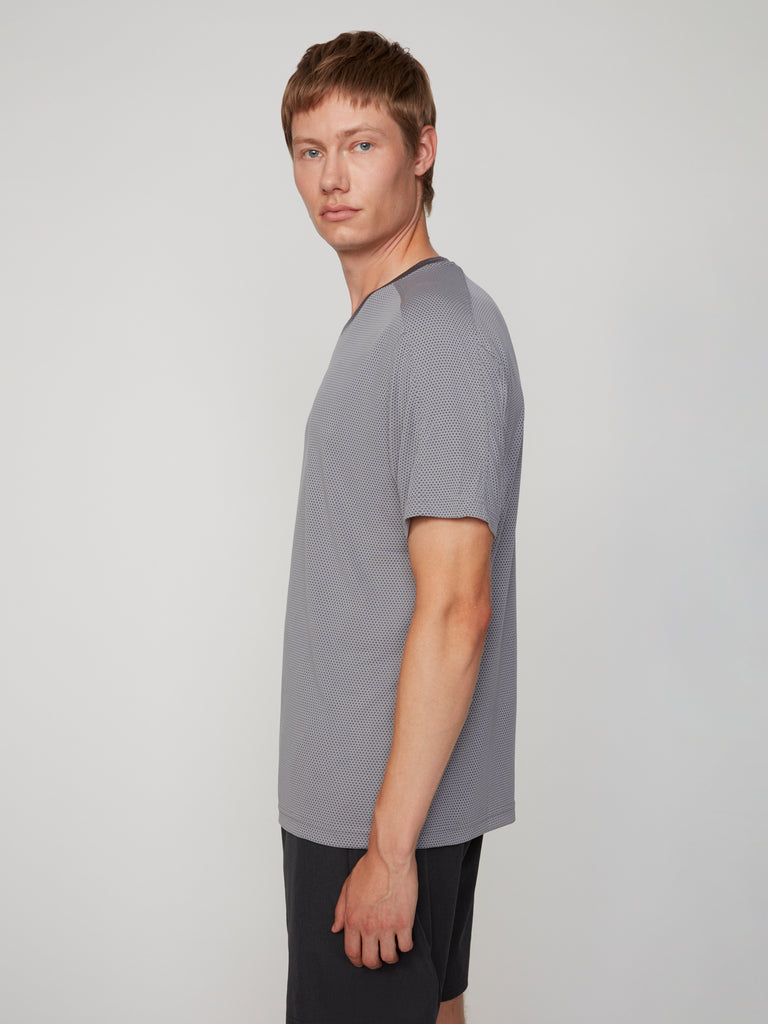 A young man with short light brown hair stands sideways, facing left, wearing the TECH APPAREL MEN’S EYELET JACQUARD CREW NECK T-SHIRT in light gray and dark shorts against a plain light gray background.