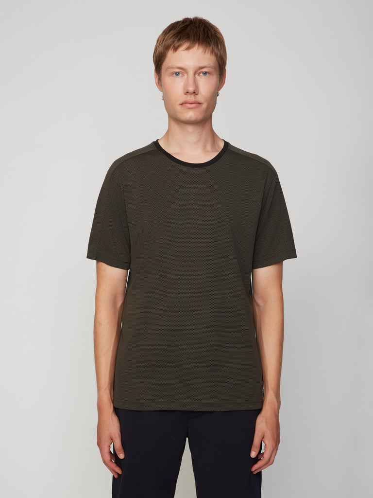 A young person with short light brown hair stands facing forward, wearing a dark green TECH APPAREL MEN’S EYELET JACQUARD CREW NECK T-SHIRT and black pants against a plain light gray background.