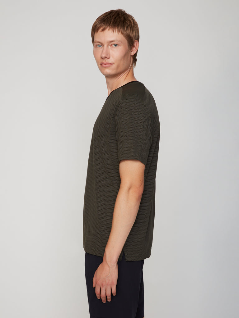 A young man with short, light brown hair stands sideways, turning his head toward the camera. He wears a dark green TECH APPAREL MEN’S EYELET JACQUARD CREW NECK T-SHIRT and black pants against a light gray background.