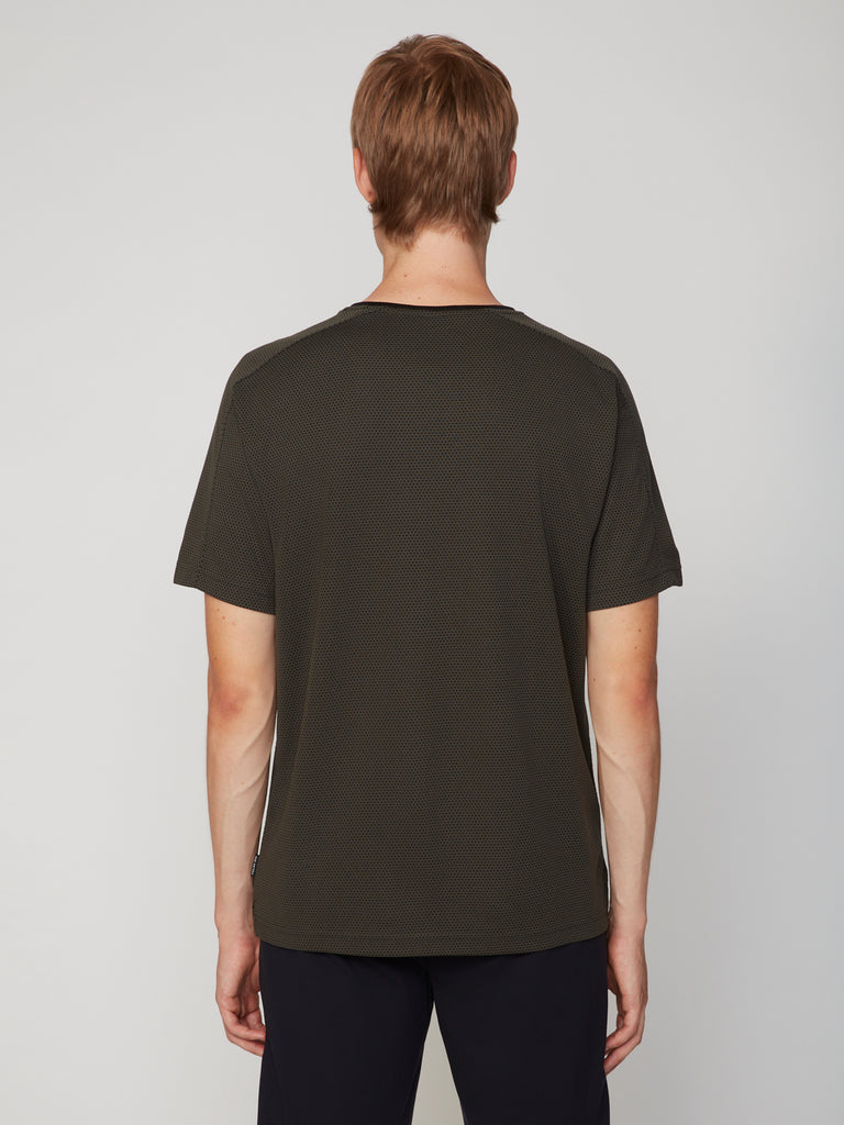 A person with short, light brown hair faces away from the camera, wearing TECH APPAREL’s MEN’S EYELET JACQUARD CREW NECK T-SHIRT and dark pants against a plain light gray background.