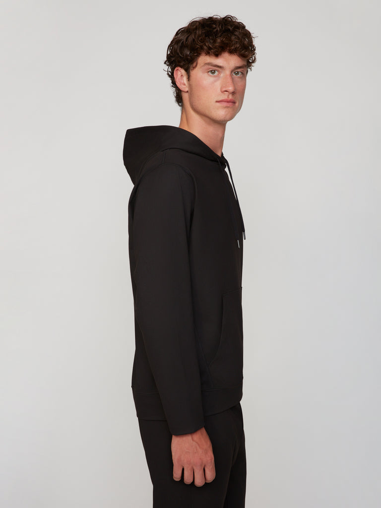 A young man with curly brown hair models the TECH APPAREL MEN’S FLEXURA FULL-ZIP MOCK NECK – LONG SLEEVE in black, standing sideways and looking at the camera against a plain light background for breathable comfort.