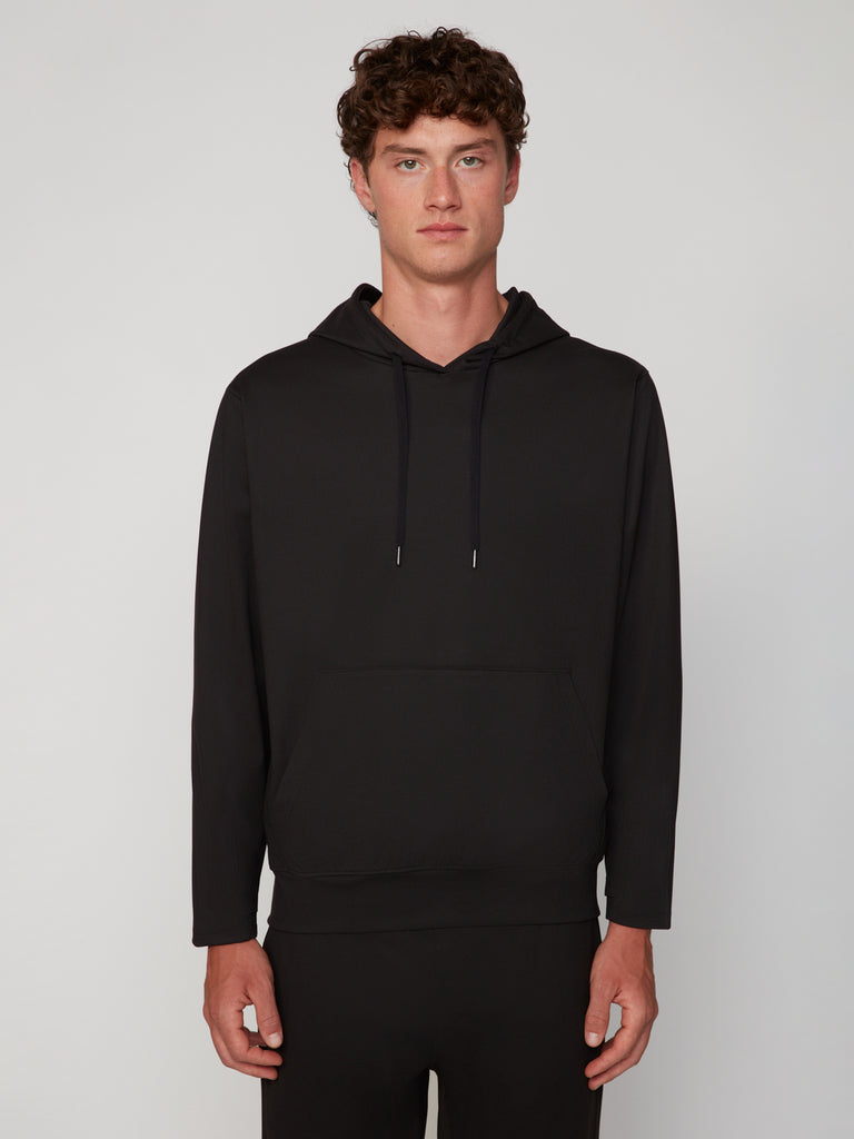 A young man with curly brown hair models the TECH APPAREL MEN’S FLEXURA FULL-ZIP MOCK NECK – LONG SLEEVE, a breathable black sweatshirt, standing against a neutral background and looking straight at the camera.