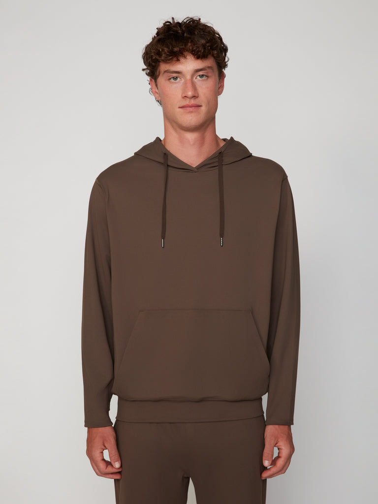 A young man with curly brown hair wears the TECH APPAREL MEN’S TECH:SOFT CLASSIC FIT HOODIE and matching pants, made from soft knit fabric, standing against a light gray background and facing forward with a neutral expression.