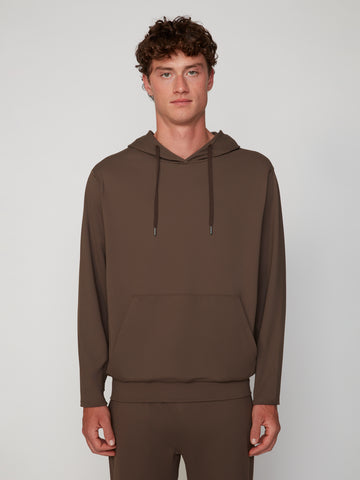 A young man with curly brown hair wears the TECH APPAREL MEN’S TECH:SOFT CLASSIC FIT HOODIE and matching pants, made from soft knit fabric, standing against a light gray background and facing forward with a neutral expression.
