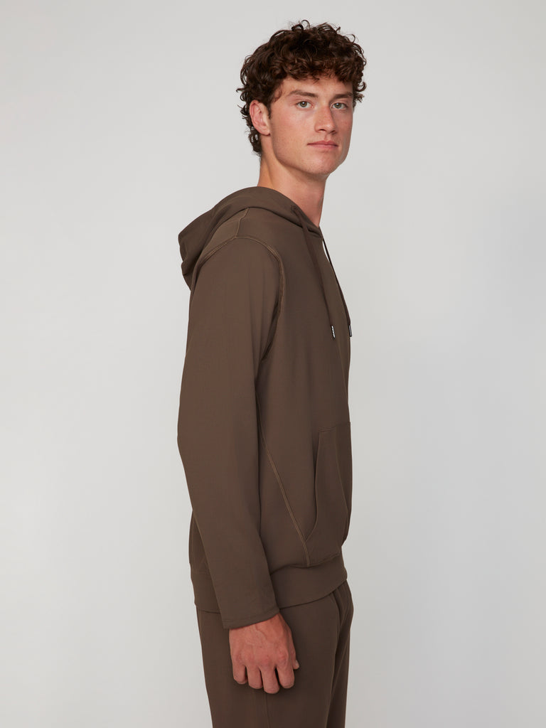 A young man with curly brown hair stands sideways in a TECH APPAREL MEN’S TECH:SOFT CLASSIC FIT HOODIE and matching pants, made from soft knit fabric for lightweight warmth, set against a plain light background.
