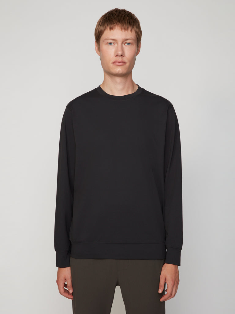 A person with short light brown hair stands against a plain light gray background, wearing the TECH APPAREL MEN’S TECH:SOFT CLASSIC FIT LONG SLEEVE PULL-OVER paired with dark pants, facing the camera with a neutral expression.