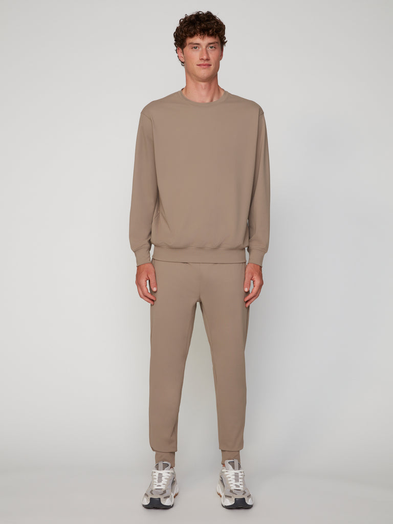 A man stands against a plain background wearing the TECH APPAREL MEN’S TECH:SOFT CLASSIC FIT LONG SLEEVE PULL-OVER in taupe with matching jogger pants, paired with white and gray sneakers for breathable warmth.