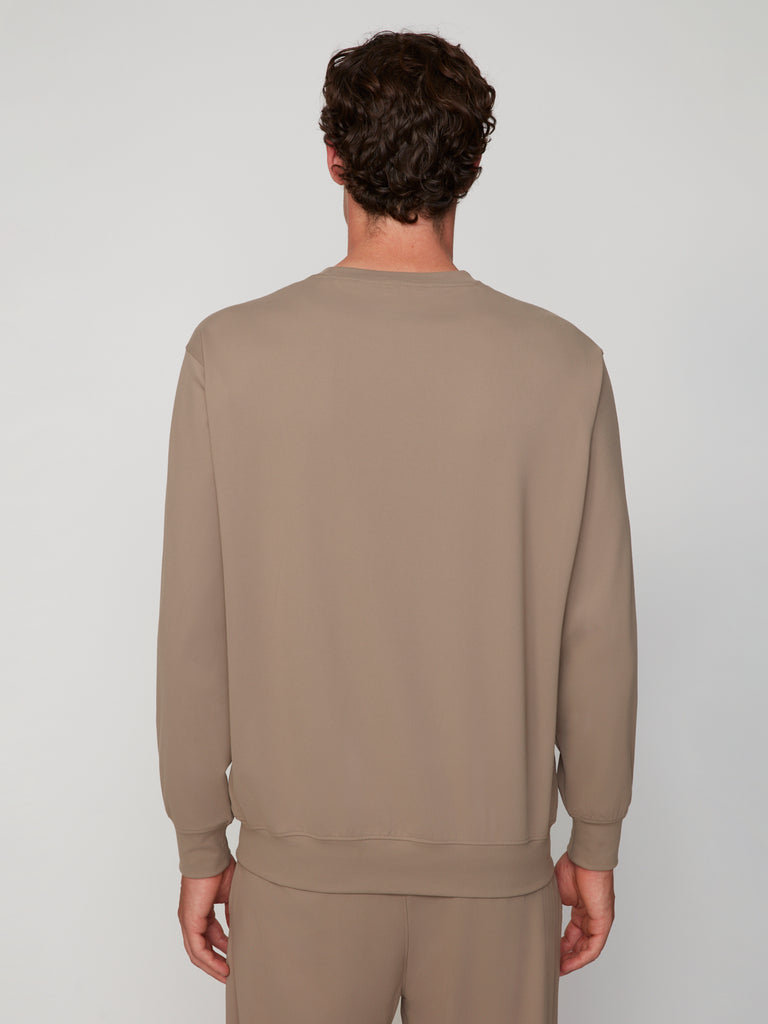 A man with short, curly brown hair is seen from behind wearing a taupe TECH APPAREL MEN’S TECH:SOFT CLASSIC FIT LONG SLEEVE PULL-OVER and matching pants, featuring breathable warmth and tech:soft fabric, against a plain light gray background.