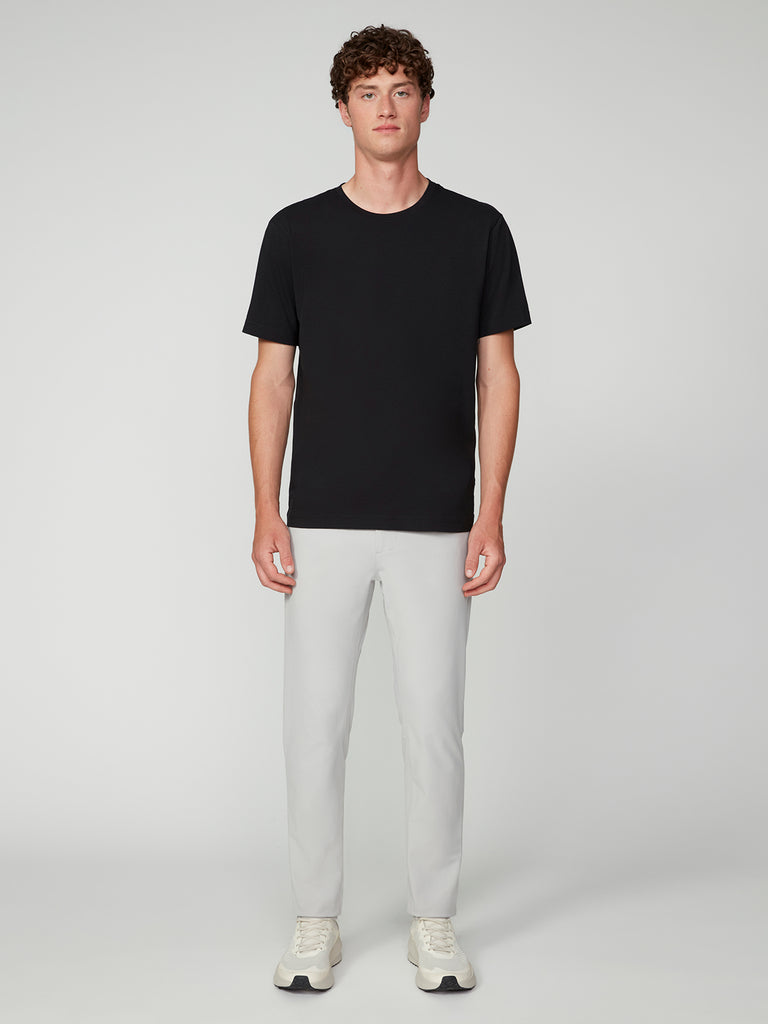 A person stands facing forward against a plain light background, wearing a black short-sleeve t-shirt, TECH APPAREL MENS FLUX 2.0 5-POCKET PANT in light gray stretch fabric, and white sneakers.