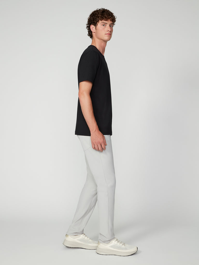 A man with curly hair stands sideways in a black t-shirt, TECH APPAREL MENS FLUX 2.0 5-POCKET PANT in light gray, and white sneakers against a plain light background.