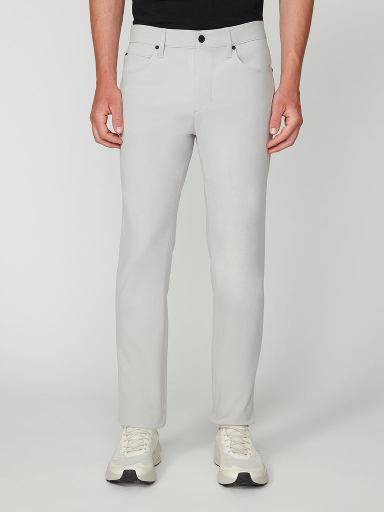 A person wears TECH APPARELs MENS FLUX 2.0 5-POCKET PANT in light gray, made from wrinkle-resistant stretch flux fabric, paired with a black shirt and white sneakers. The image is cropped at the shoulders against a plain white background.