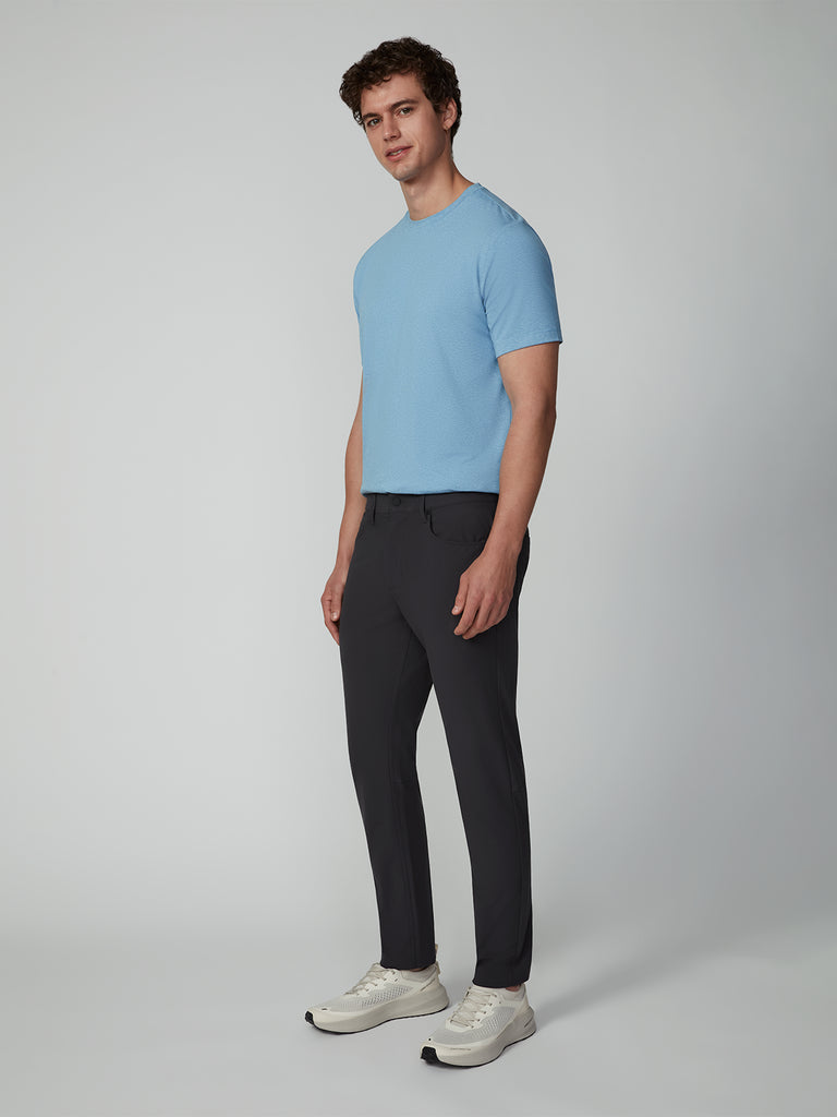 A man stands against a plain light background, wearing a light blue short-sleeve shirt, TECH APPAREL MENS FLUX 2.0 5-POCKET PANT made from stretch flux fabric, and white sneakers, looking at the camera with a neutral expression.