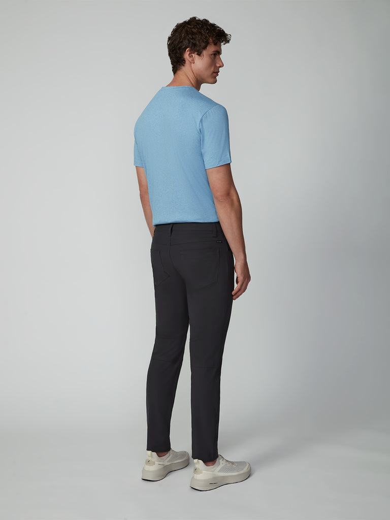 A man stands facing away, looking over his shoulder. He wears a light blue short-sleeve shirt, TECH APPAREL MENS FLUX 2.0 5-POCKET PANT in stretch flux fabric, and white sneakers against a plain light grey background.