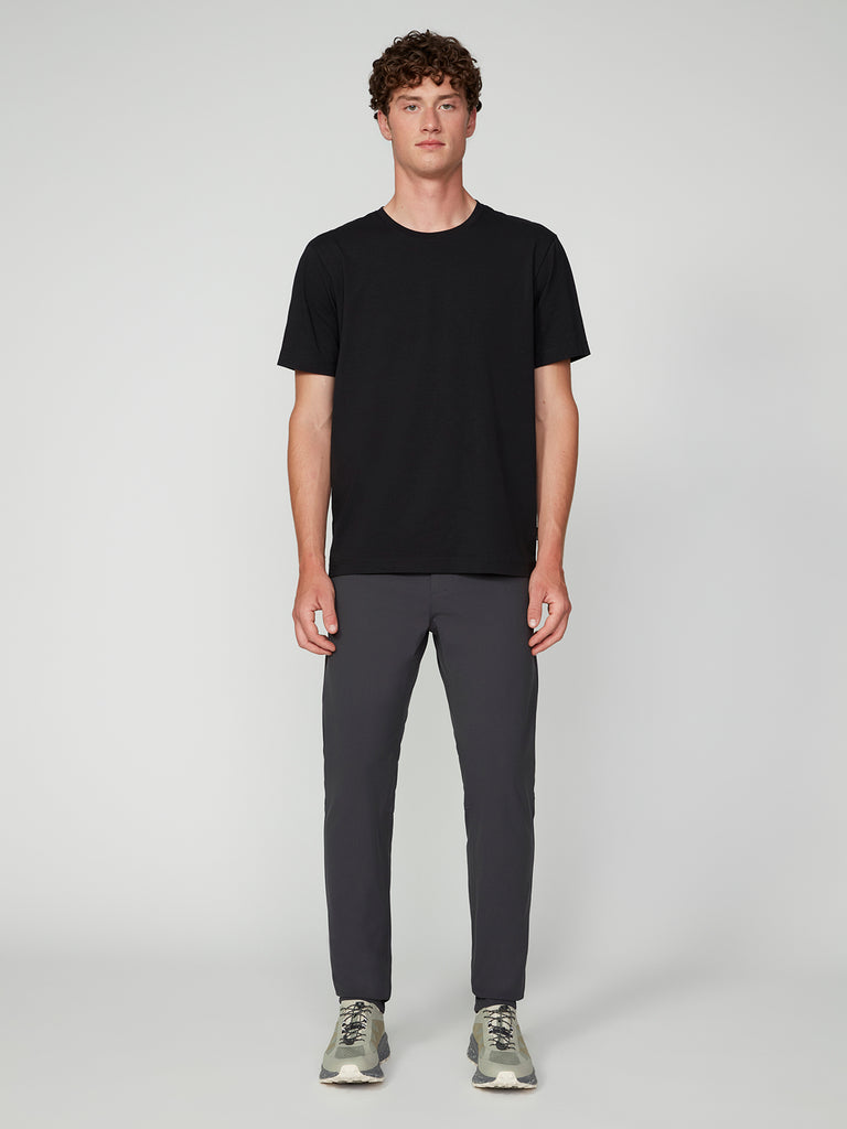 A man stands facing forward against a plain light gray background, wearing a black short-sleeve t-shirt, TECH APPAREL MENS FLUX 2.0 5-POCKET PANT in dark gray, and light gray sneakers.