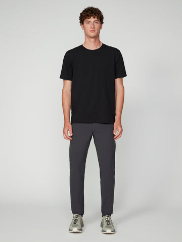 A man stands facing forward against a plain light gray background, wearing a black short-sleeve t-shirt, TECH APPAREL MENS FLUX 2.0 5-POCKET PANT in dark gray, and light gray sneakers.