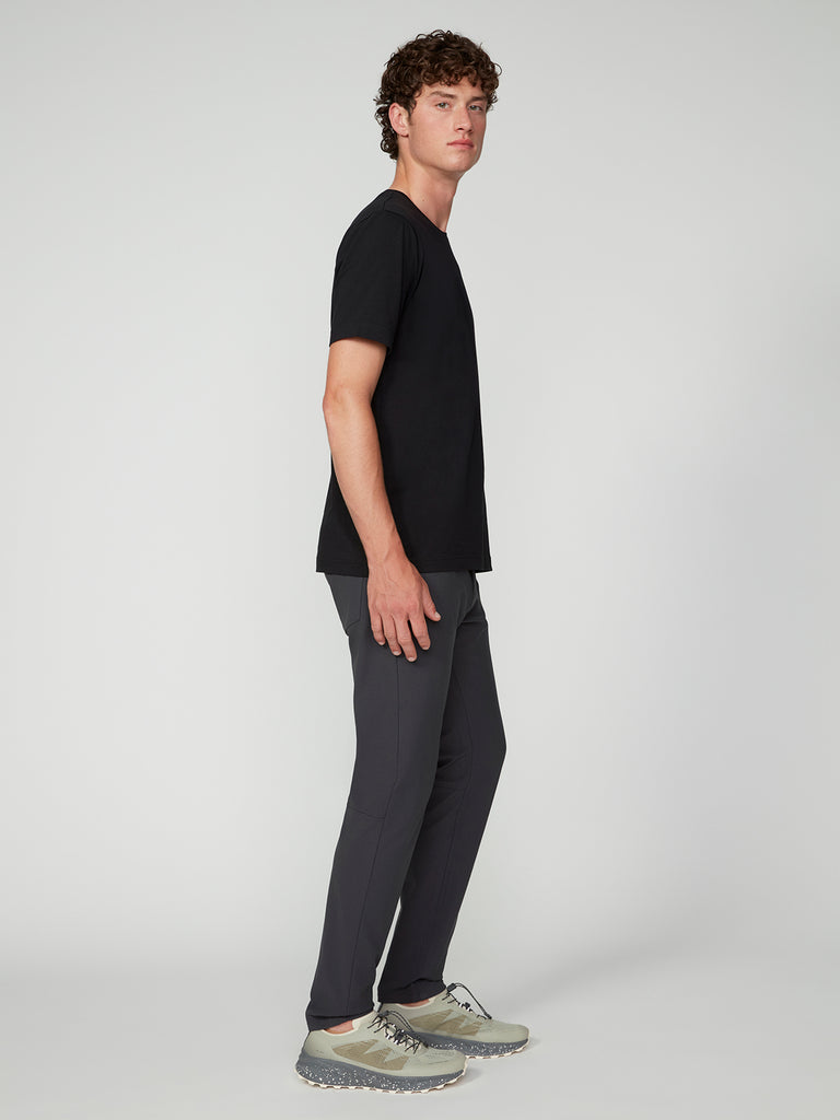 A young man with short curly hair stands sideways, facing the camera. Hes wearing a black t-shirt, TECH APPAREL MENS FLUX 2.0 5-POCKET PANT in stretch flux fabric, and gray sneakers against a plain light background.