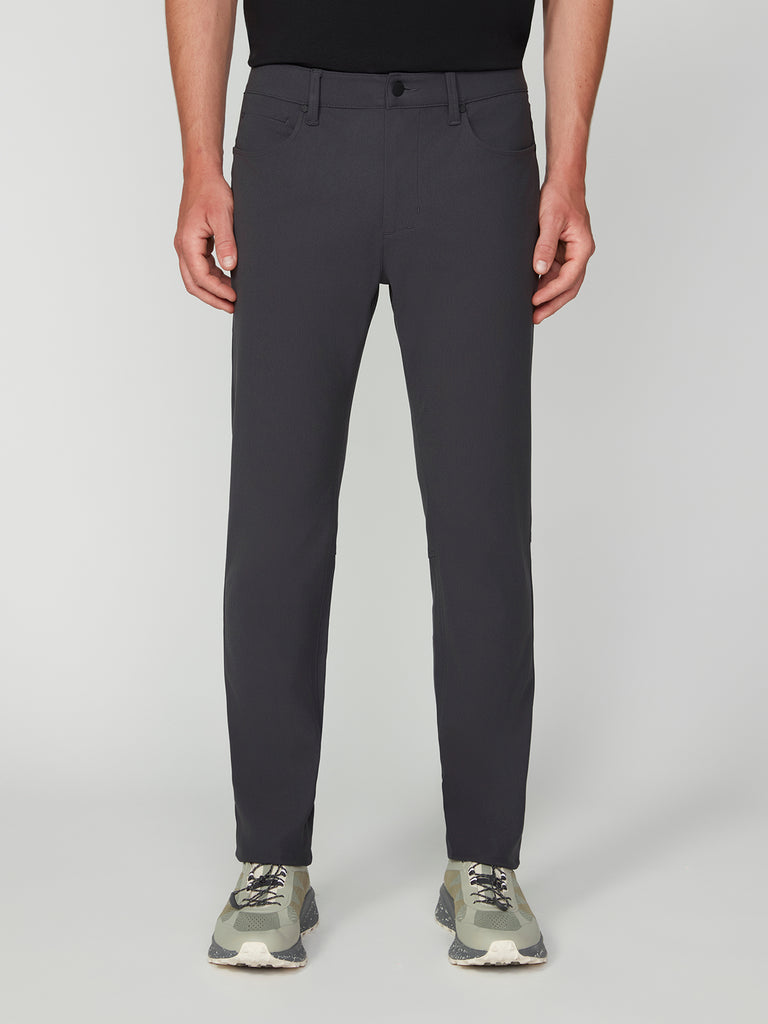 A person wearing TECH APPAREL MENS FLUX 2.0 5-POCKET PANT, a black shirt, and gray-green sneakers stands against a plain light background. The photo is cropped from shoulders to feet.