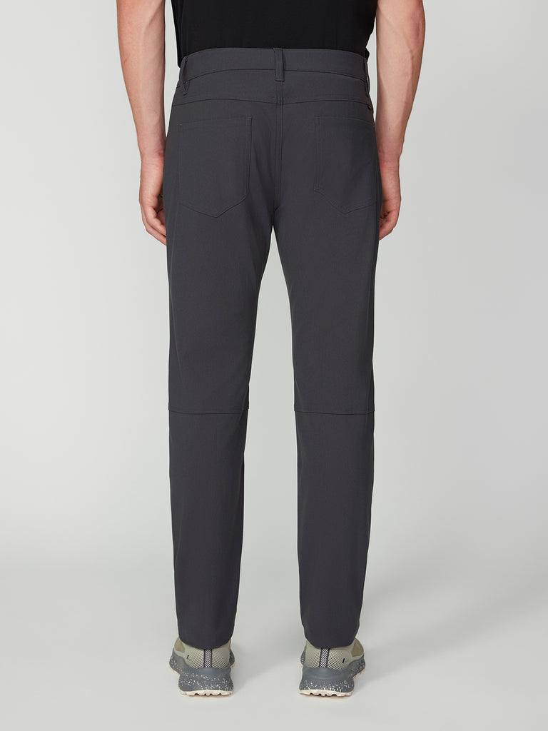 A person is shown from the back in TECH APPAREL MENS FLUX 2.0 5-POCKET PANT, dark gray and wrinkle-resistant with back pockets and knee seams, paired with gray sneakers on a light gray background.