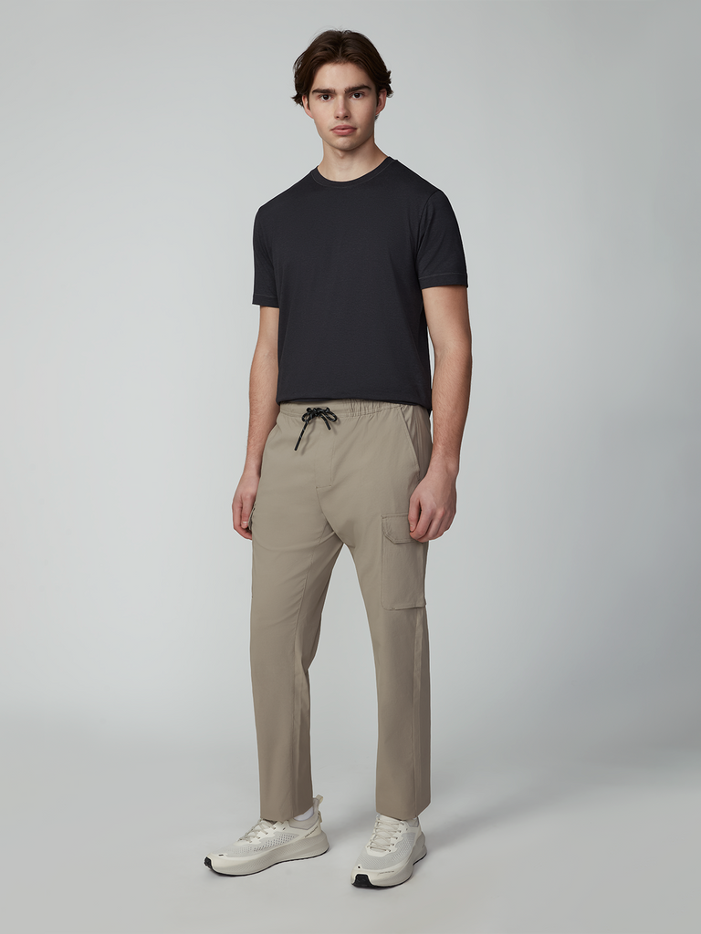 A young man in a black tee and white sneakers models TECH APPARELs MENS FLUX 2.0 5-POCKET PANT—beige, wrinkle-resistant, and made from stretch flux fabric—standing against a plain background with a neutral look.