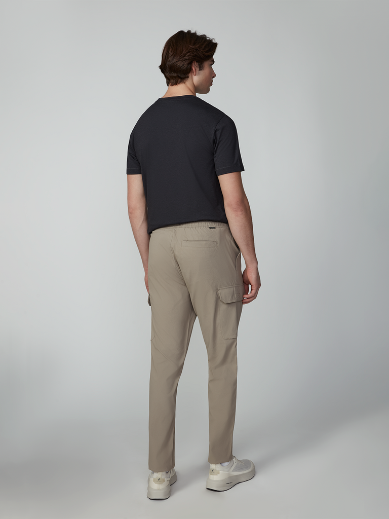 A man faces away from the camera, wearing a black short-sleeve t-shirt, TECH APPAREL MENS FLUX 2.0 5-POCKET PANT in beige (wrinkle-resistant, straight cut), and white sneakers against a plain light gray background.