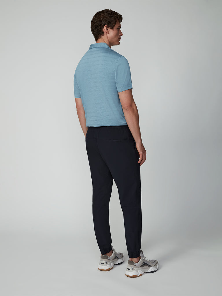 A man faces away from the camera wearing a light blue short-sleeve polo, TECH APPAREL MENS FLUX 2.0 JOGGER pants, and gray sneakers, standing against a plain light gray background.