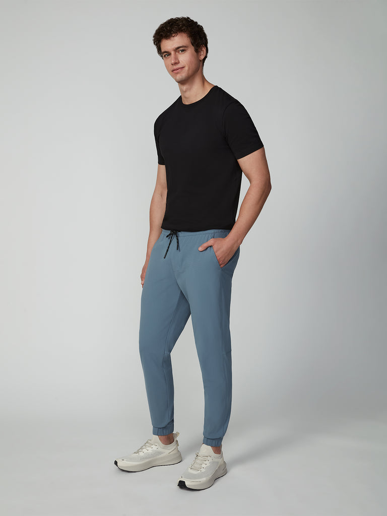 A man stands in a studio wearing a black t-shirt and TECH APPAREL MENS FLUX 2.0 JOGGER in light blue, made with stretch flux fabric. He faces slightly toward the camera, hands in pockets, set against a plain light background.