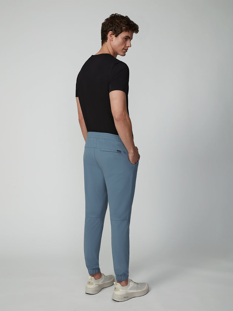 A man stands facing away, looking over his shoulder. He wears a black t-shirt, TECH APPAREL MENS FLUX 2.0 JOGGER in light blue, and white sneakers against a plain, light-colored background.