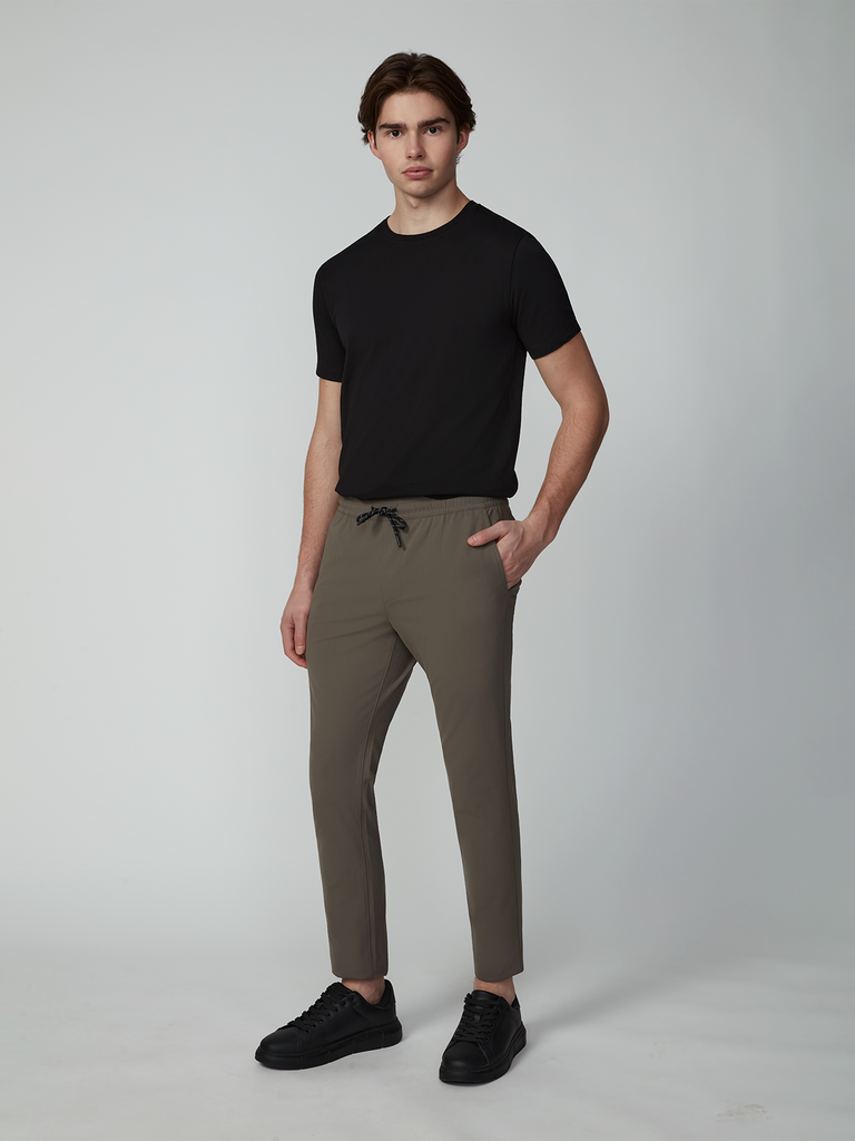 A young man faces the camera with a neutral expression, wearing TECH APPAREL MENS FLUX 2.0 PULL-ON PANT, a black T-shirt, and black sneakers; one hand is in his pocket against a plain background.