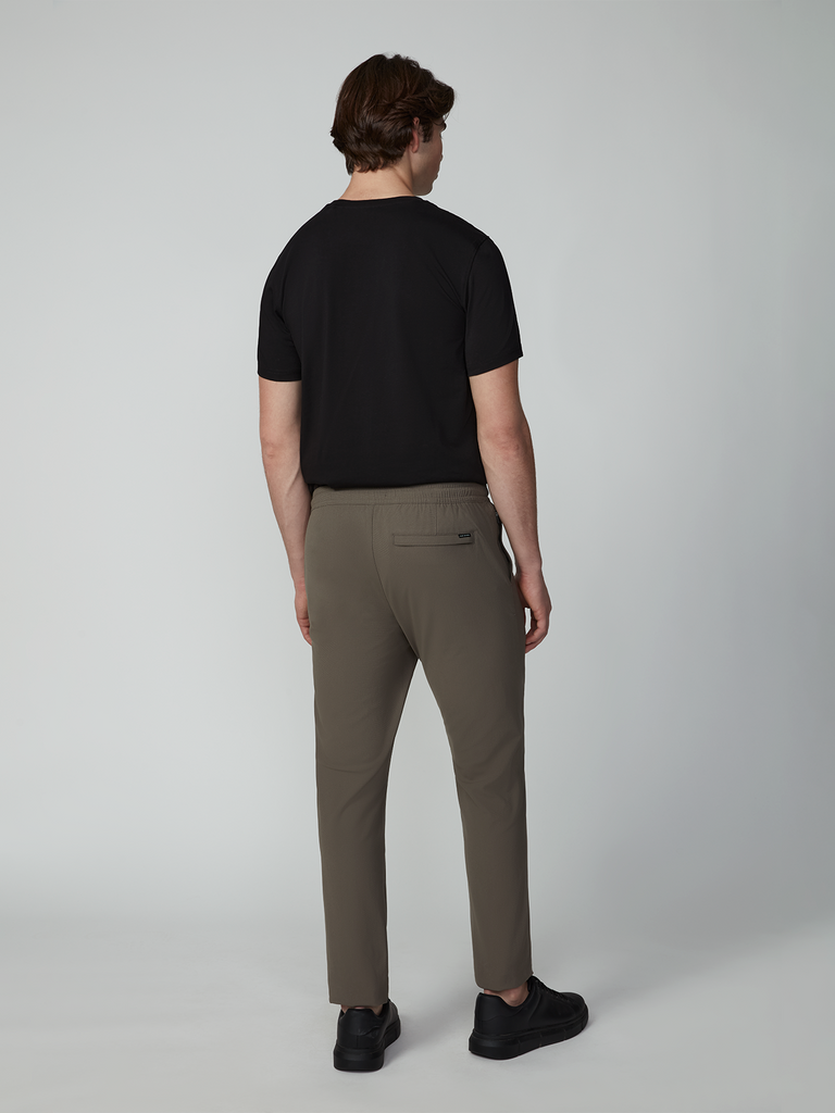 A man with short brown hair faces away, wearing a black T-shirt and TECH APPAREL MENS FLUX 2.0 PULL-ON PANT in olive green, paired with black shoes against a plain light background.