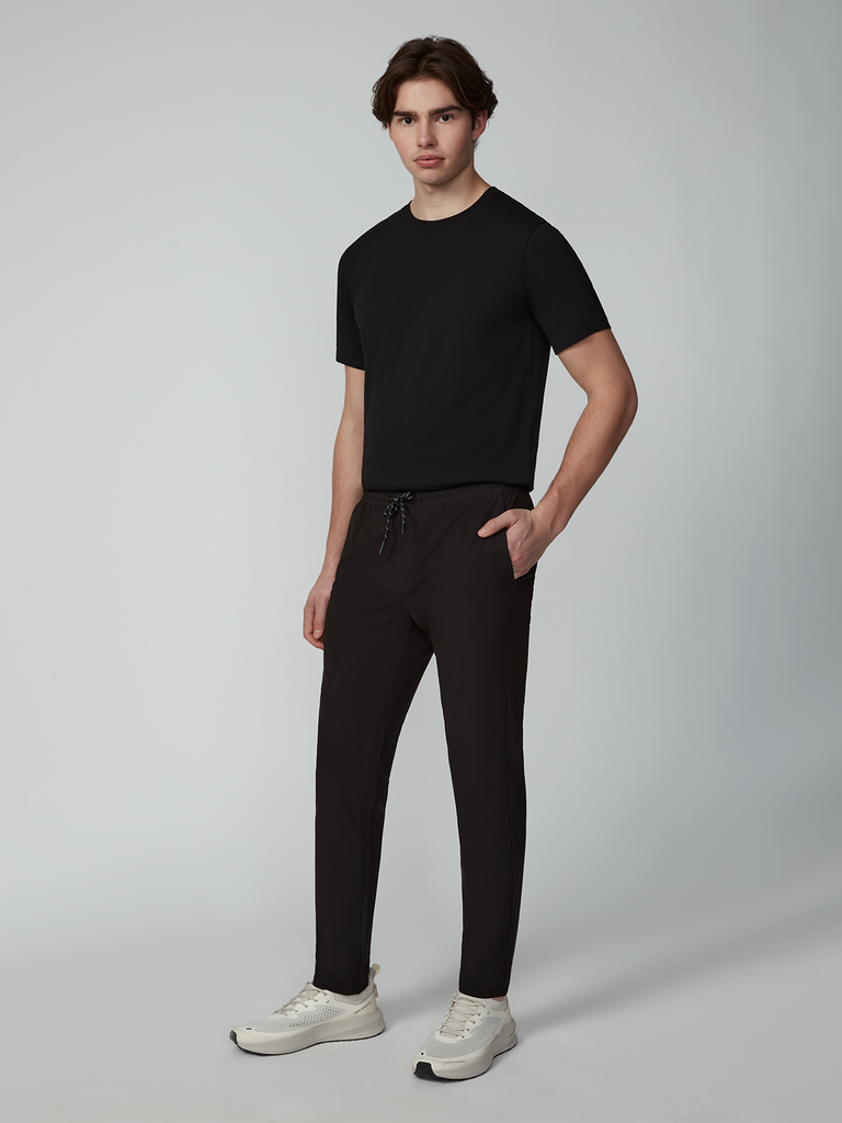 A man stands against a plain background wearing a black T-shirt, TECH APPAREL MENS FLUX 2.0 PULL-ON PANT in black, and white sneakers. With one hand in his pocket, he looks towards the camera.