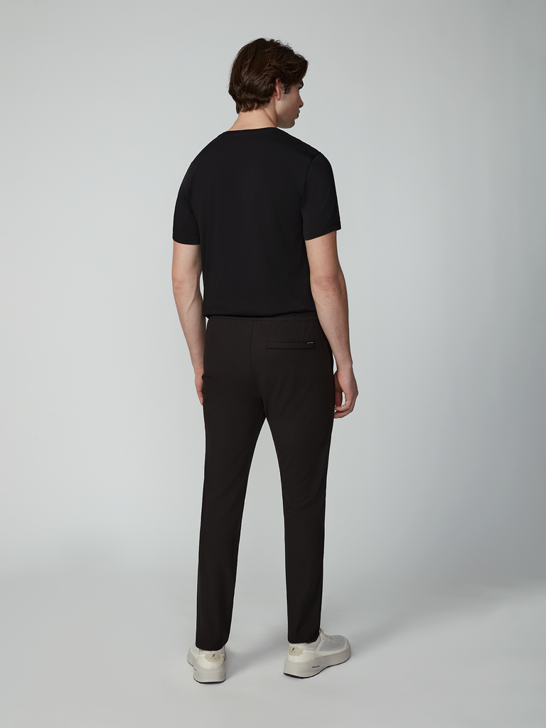 A man with brown hair stands facing away from the camera, wearing a black wrinkle-resistant short-sleeve shirt, TECH APPAREL MENS FLUX 2.0 PULL-ON PANT, and white sneakers, against a plain light gray background.