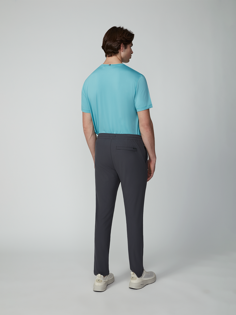 A man faces away, wearing TECH APPARELs MENS FLUX 2.0 PULL-ON PANT with a drawcord waist, a light blue t-shirt, and white sneakers against a plain light gray background.